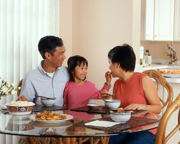 Asian family sits around the table for lunch while young girl offers her mother a bite of food